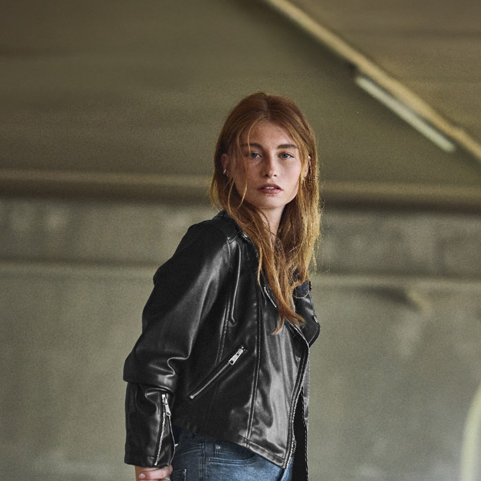 Vrouw met lang rood haar draagt een zwart leren jack en jeans.