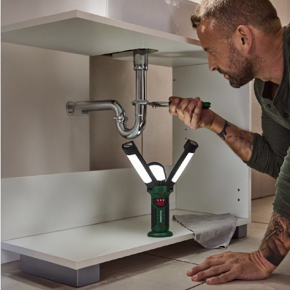 Man repareert gootsteenafvoer met een Parkside werklamp die de ruimte verlicht.