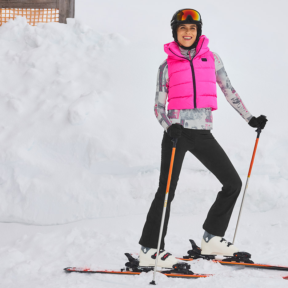 Vrouw in roze ski-bodywarmer, ski-helm en skibroek met ski's en stokken in de sneeuw.