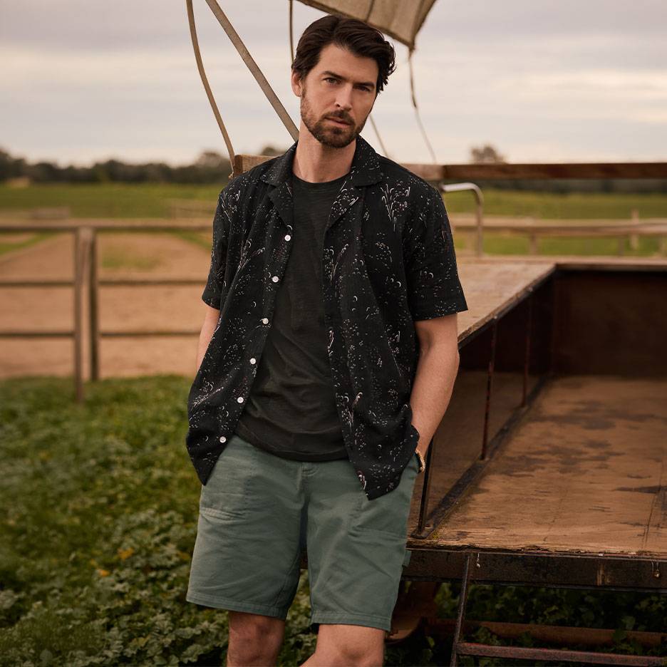 Man in zwart bedrukt overhemd, T-shirt en groene korte broek, staand bij een aanhanger.