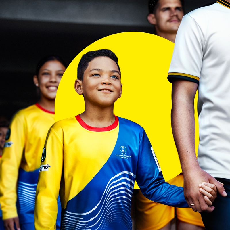 Een jonge jongen in een geel en blauw voetbalshirt houdt de hand van een volwassene vast.