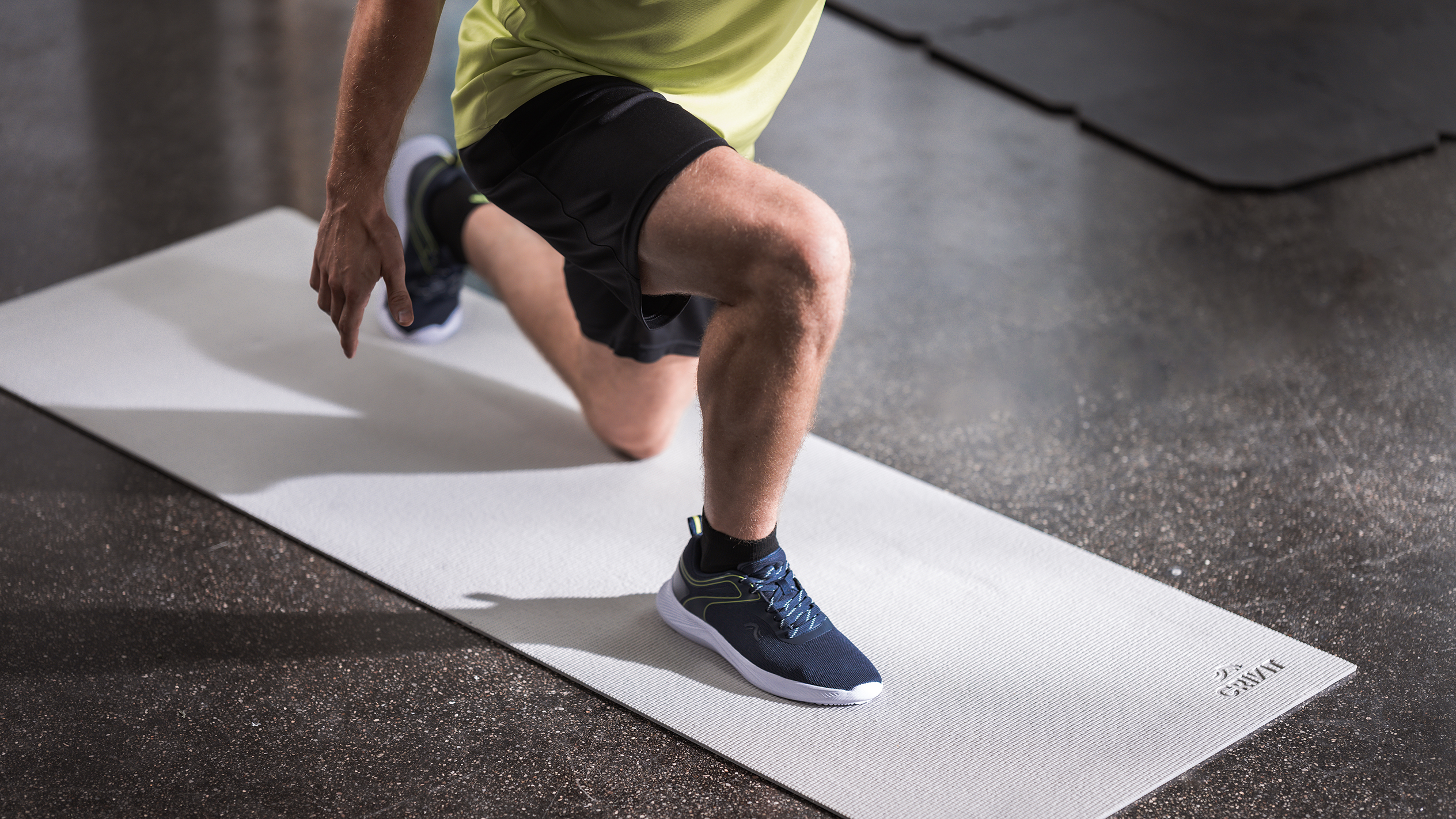 Man in sportkleding doet lunges op een lichtgrijze oefenmat.