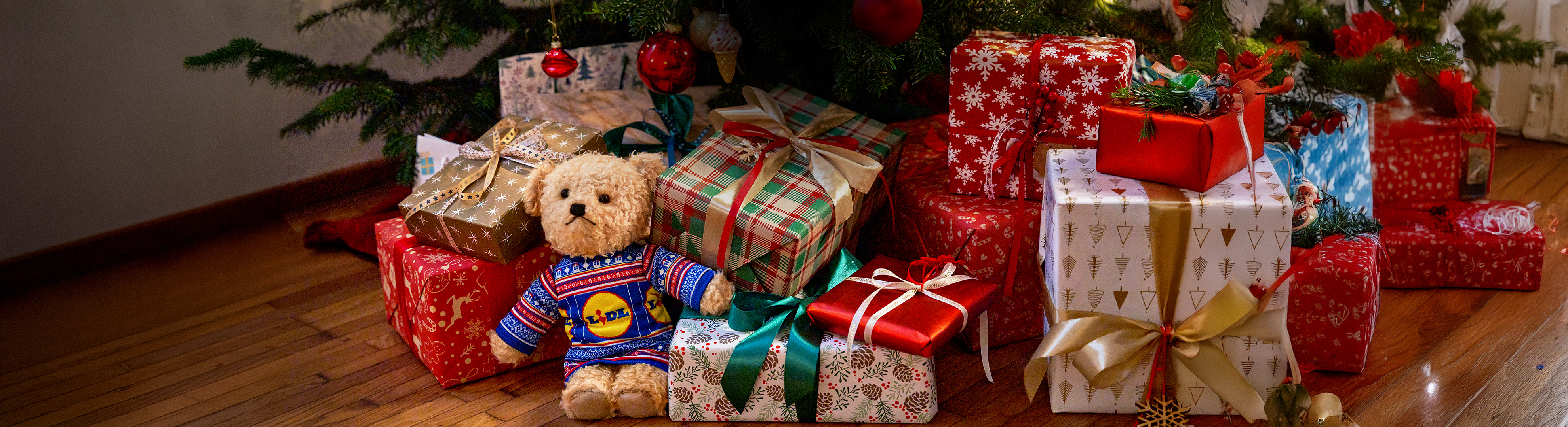 Teddybeer in Lidl-trui tussen kerstcadeaus onder een kerstboom.