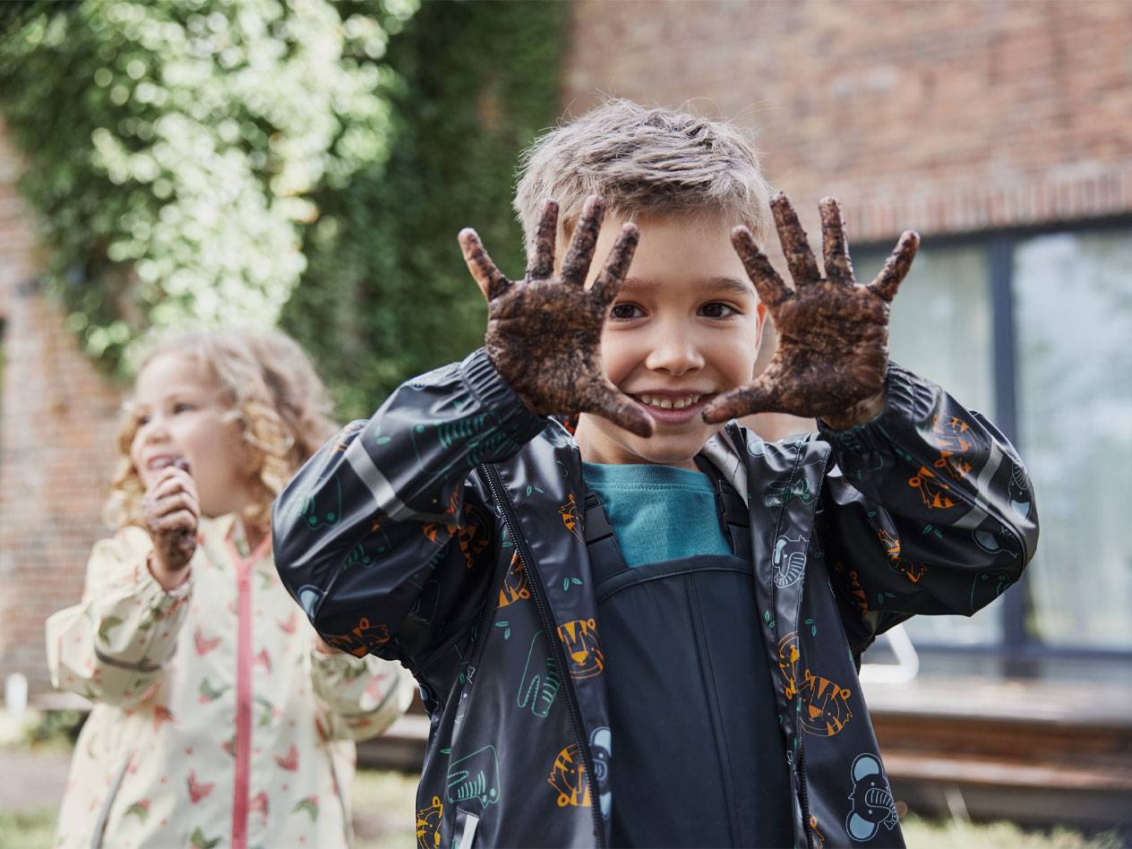 Twee kinderen in regenjassen, met modderige handen, glimlachen buiten.
