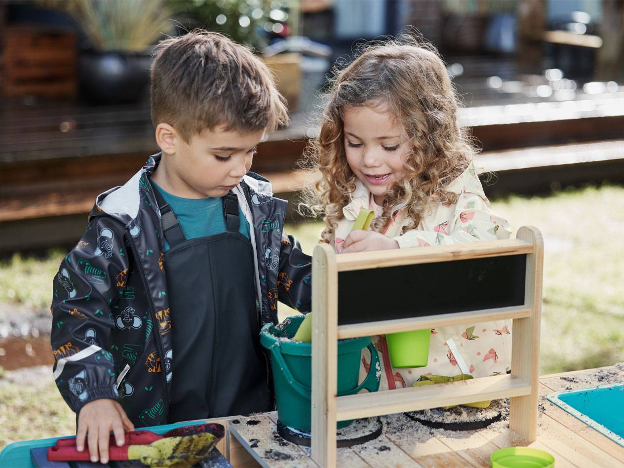 Twee kinderen in regenjassen en tuinbroeken spelen met zand aan een speeltafel.