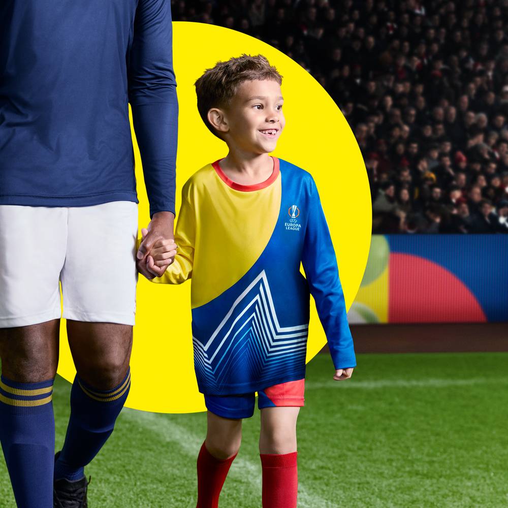 Een kind in een geel en blauw voetbalshirt met Europa League-logo, hand in hand met een volwassen speler op een voetbalveld.