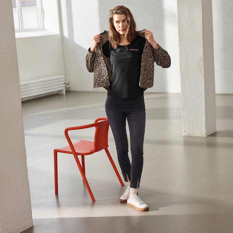 Vrouw in een zwart T-shirt met tekst, jeans en een luipaardprint jasje, staand naast een rode stoel.