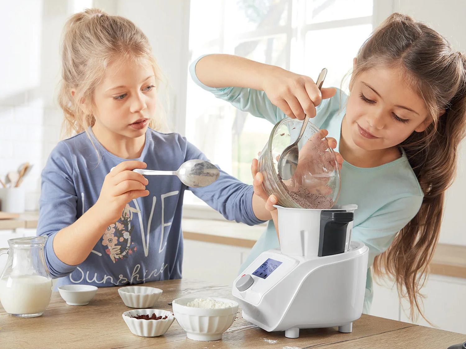 Twee meisjes bereiden een maaltijd met een keukenmachine en ingrediënten op een houten tafel.
