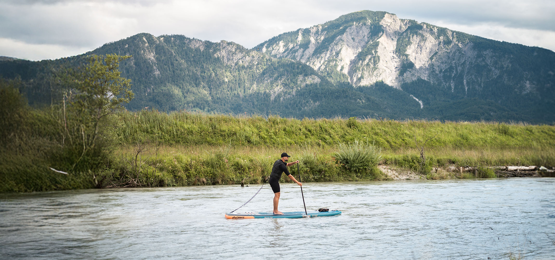 Man op paddleboard op een rivier met bergen en weelderige begroeiing op de achtergrond.