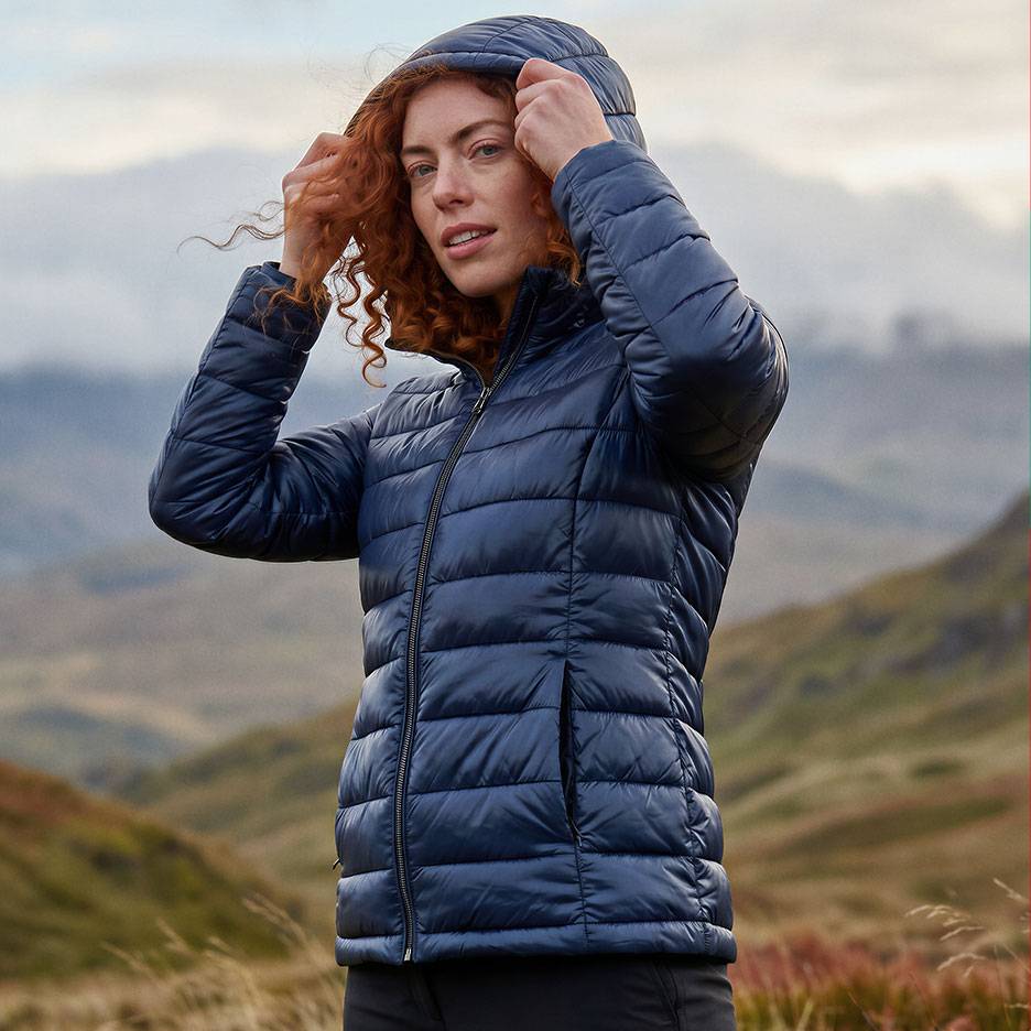 Vrouw met rood haar in een donkerblauwe gewatteerde jas met capuchon in een berglandschap.