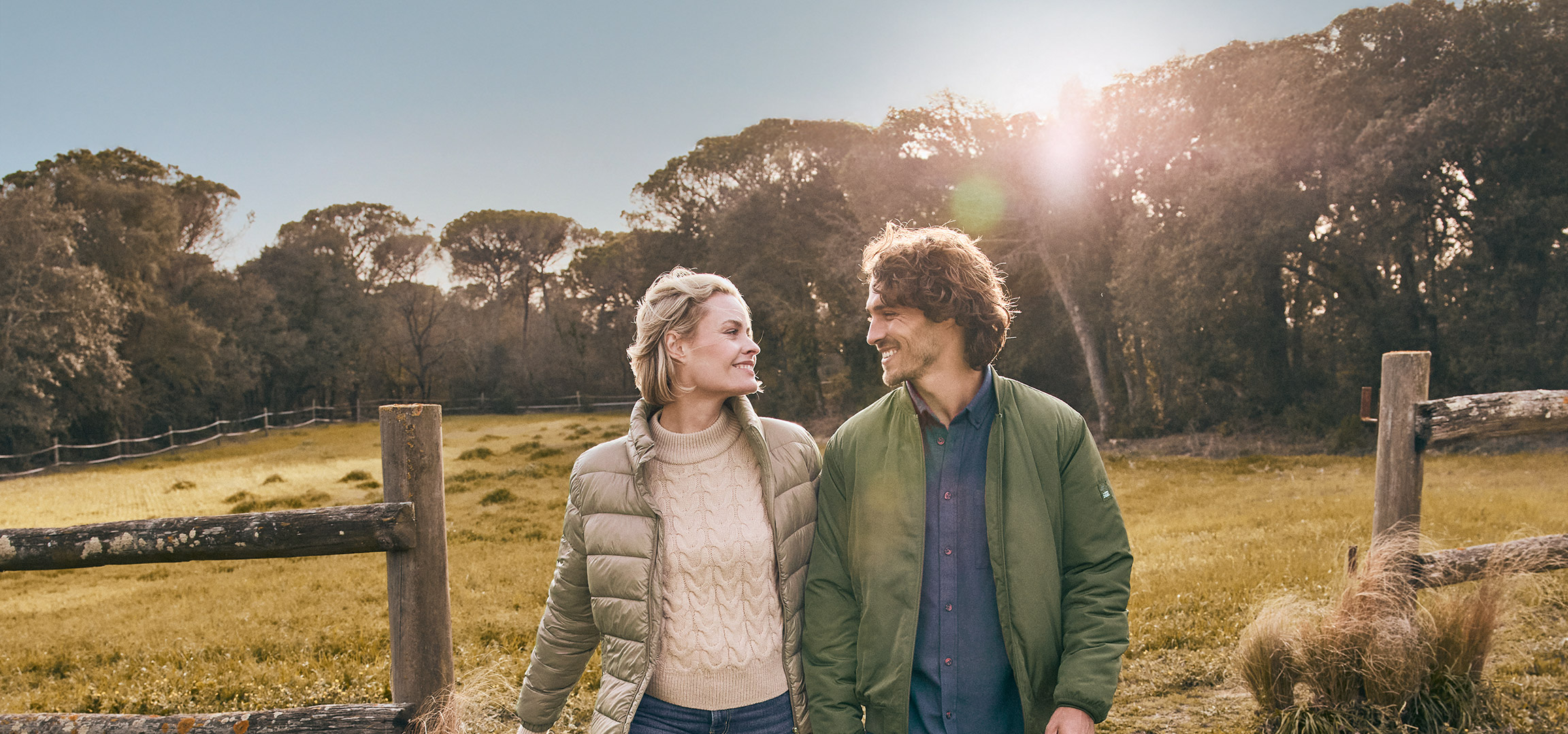 Een man en vrouw in jassen en truien, lachend in een veld met een houten hek.
