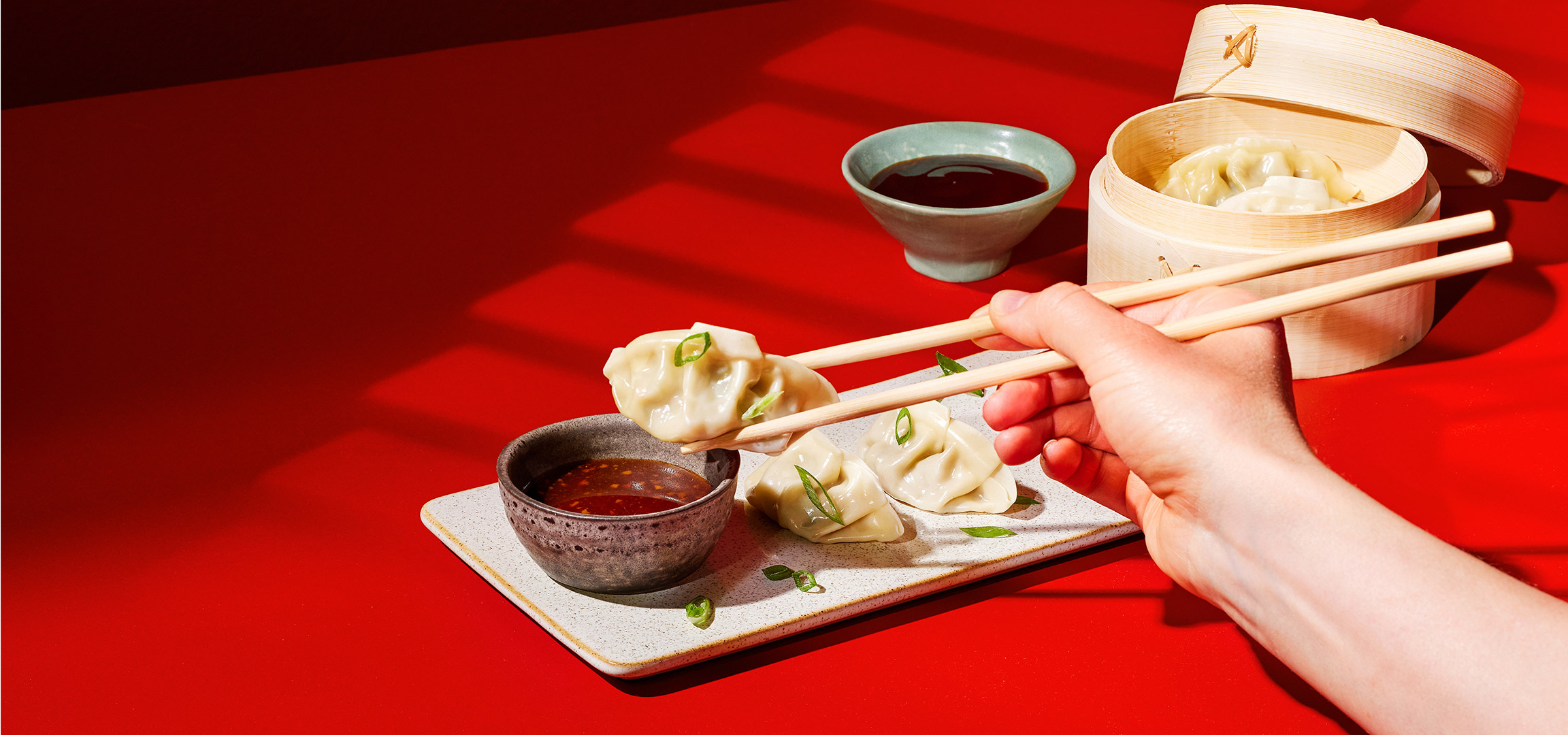 Een hand die een dumpling met eetstokjes boven een kommetje saus houdt, met meer dumplings op een schaal.