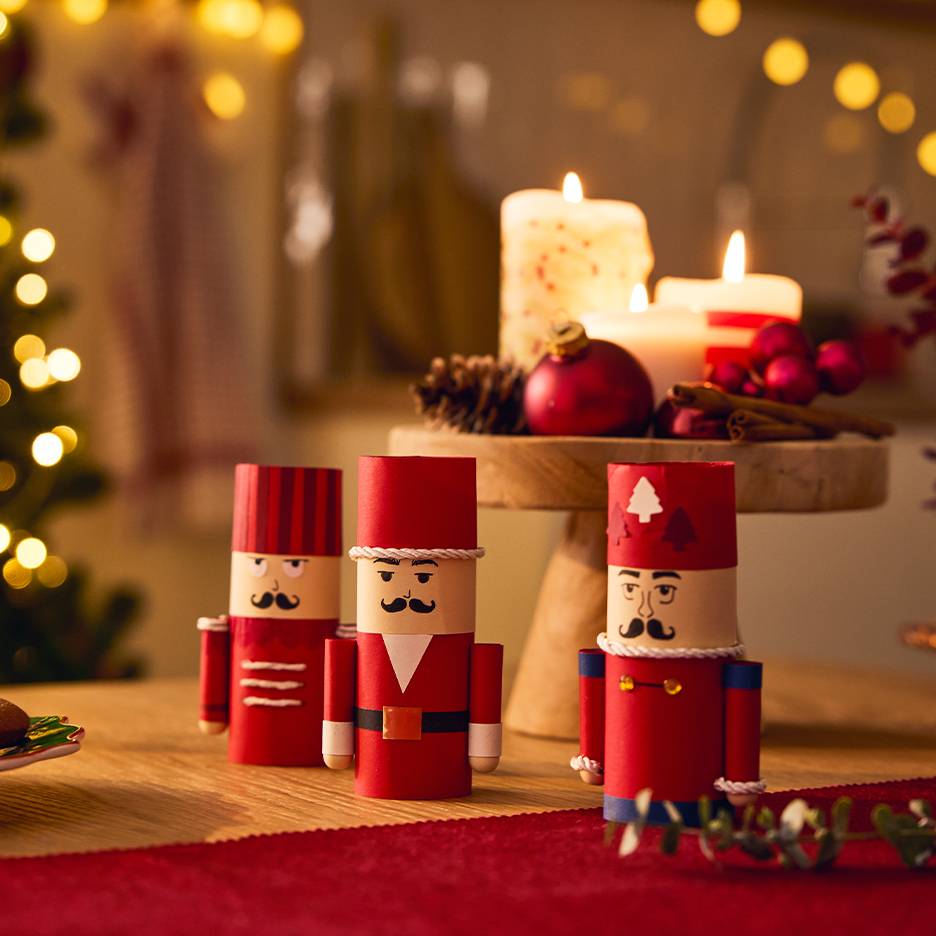 Drie feestelijke papieren notenkrakers op een tafel met kerstdecoraties en brandende kaarsen.