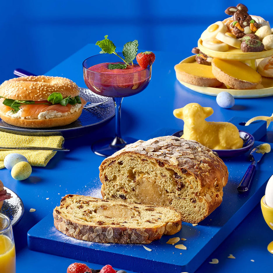 Paasontbijt met bagel, smoothie, paasbrood, paaslammetje en chocolade paaseitjes op een blauwe tafel.