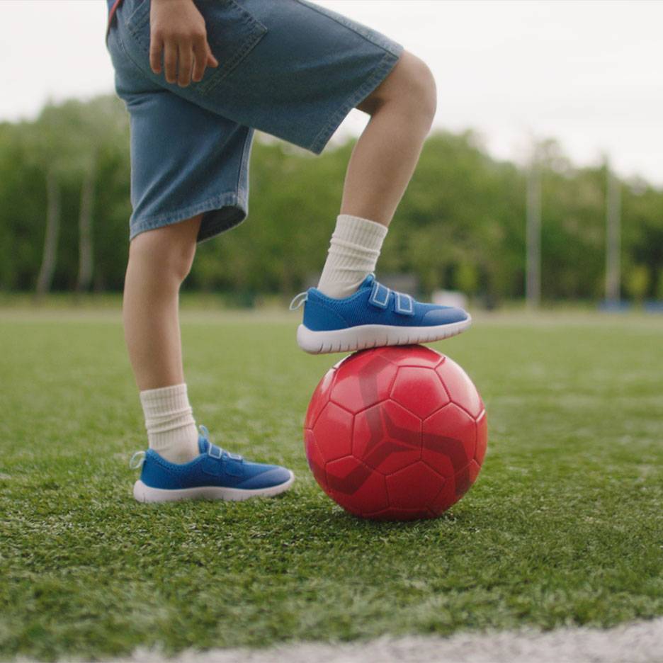 Kind in korte broek en blauwe sneakers, met één voet op een rode voetbal.