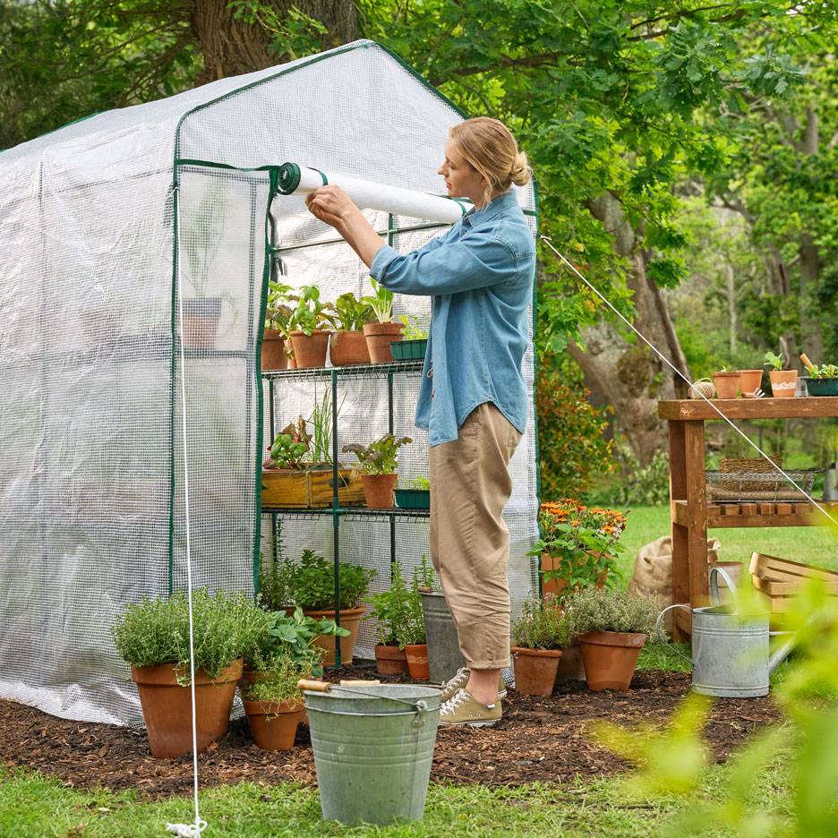 Vrouw verzorgt planten in een inloopkas met planken vol potkruiden en groenten.