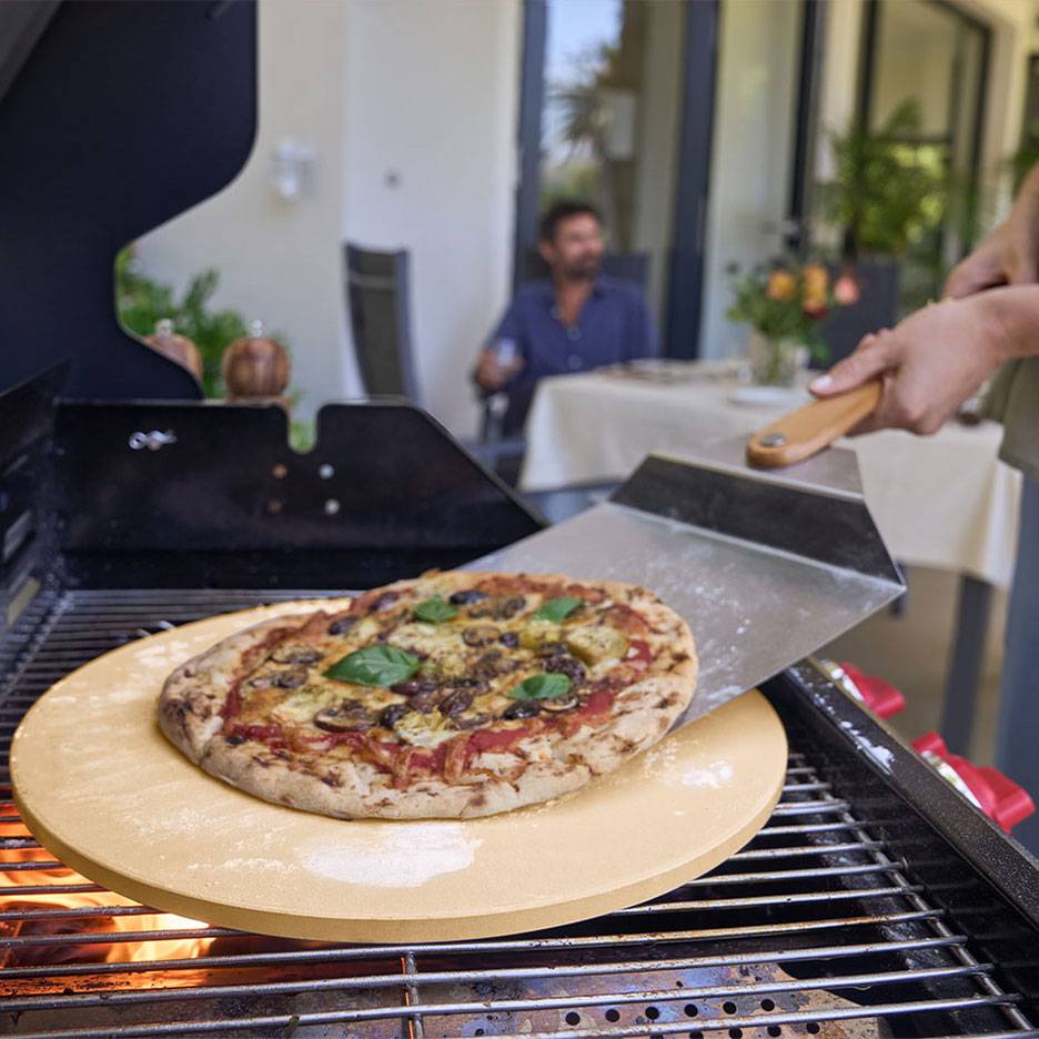 Pizza op een pizzasteen op de grill, met een pizzaschep en een man op de achtergrond.
