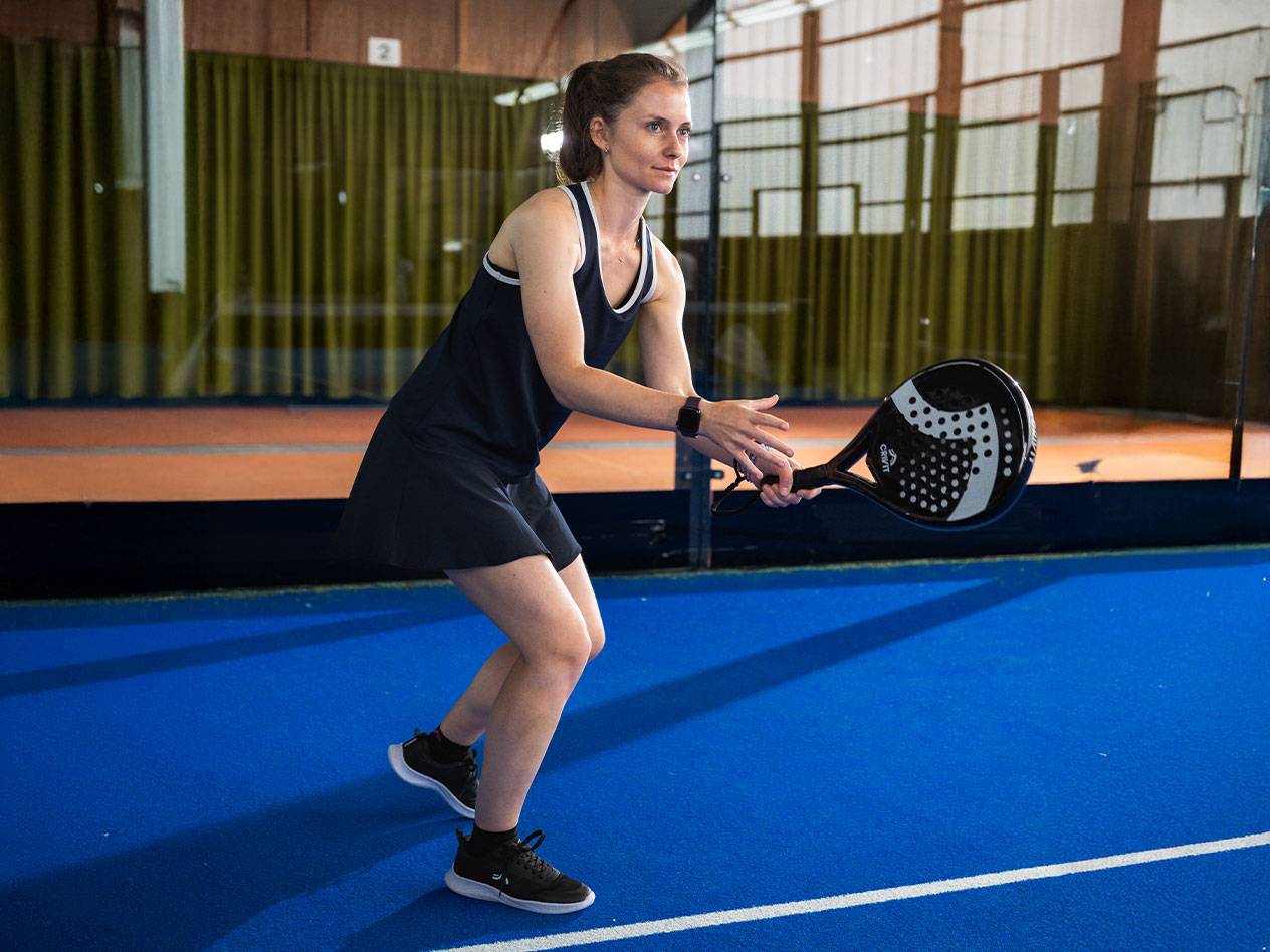 Vrouw in zwarte padeljurk met zwart padelracket op blauw veld.