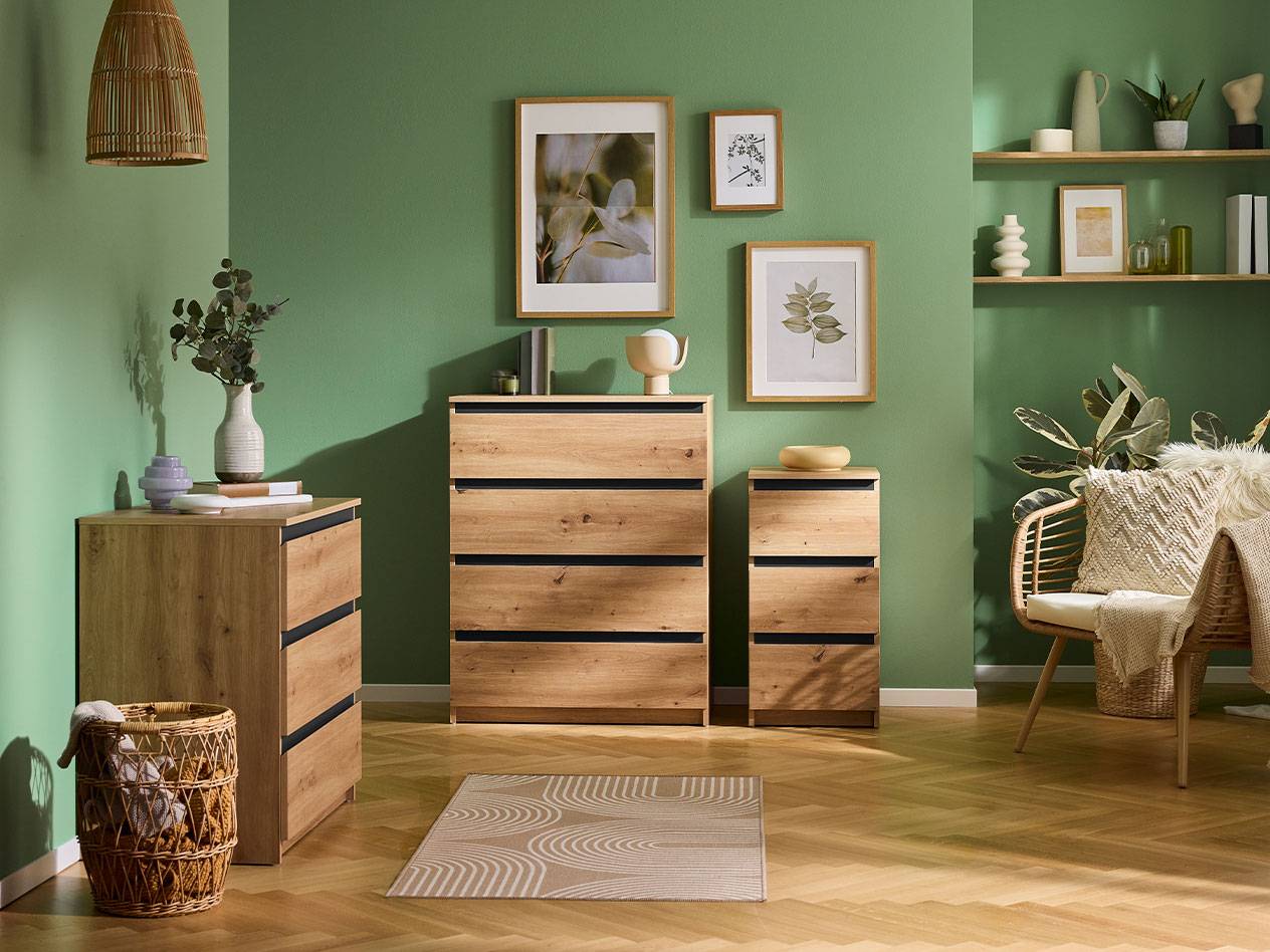 Woonkamer met groene muren, houten ladekasten, planken en een rotan fauteuil.