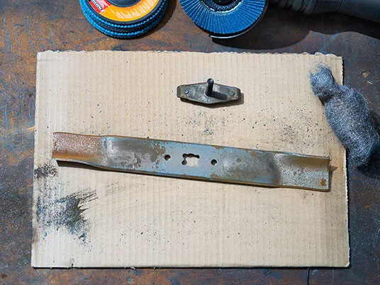 Roestig grasmaaierblad op karton met schuurschijven en staalwol.