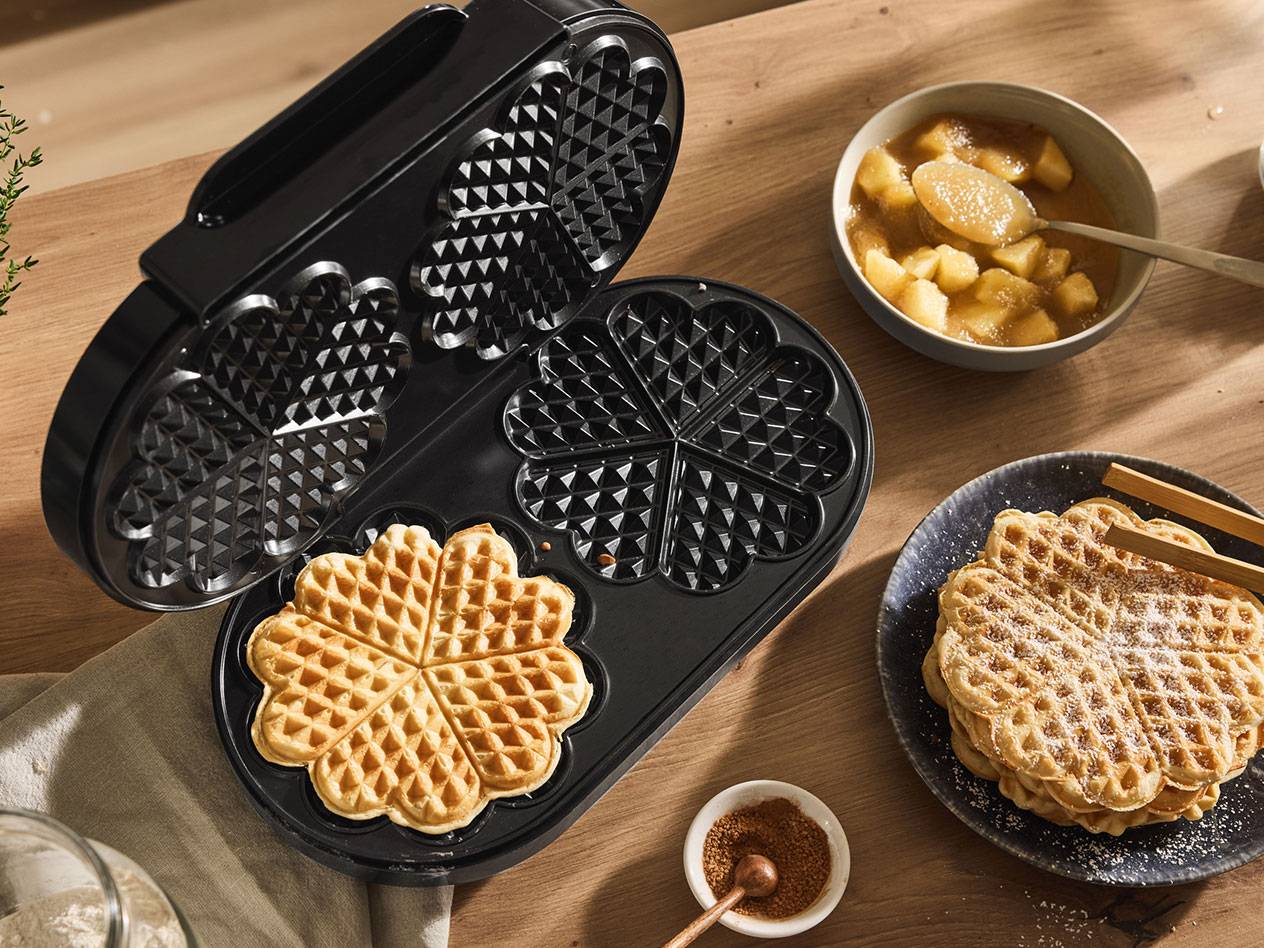 Een open wafelijzer met een hartvormige wafel, naast gebakken wafels en appelcompote.