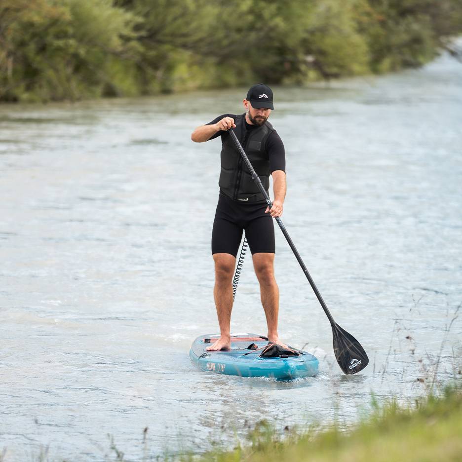 Man aan het paddleboarden op een rivier, draagt een zwart wetsuit, zwemvest en pet.