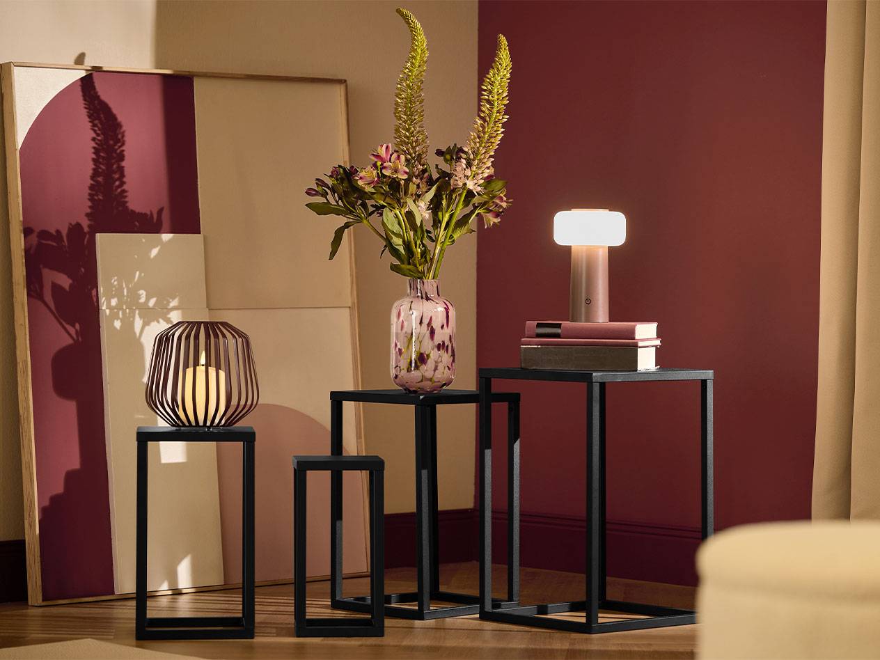 Drie zwarte bijzettafels met een kaars, een vaas bloemen en een lamp.