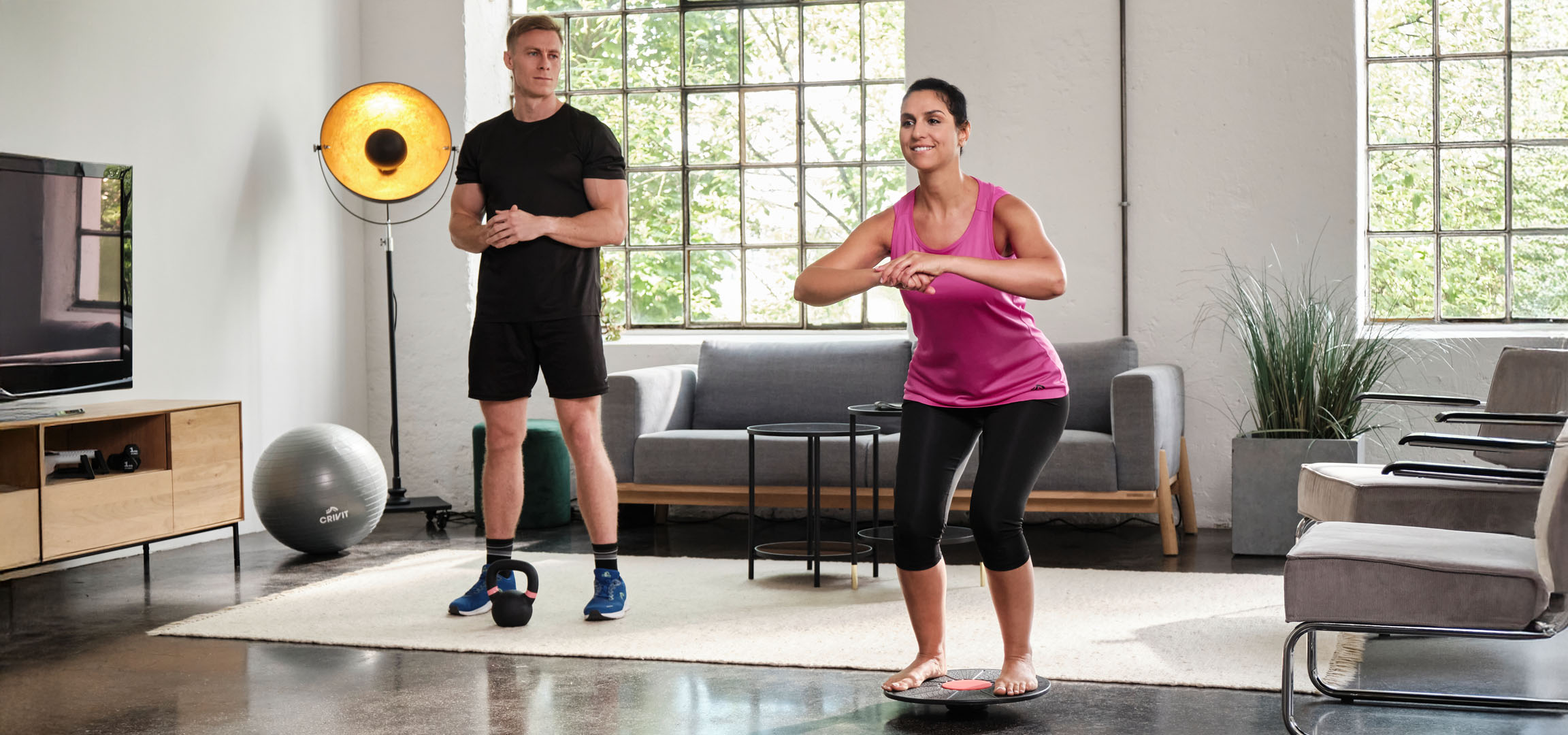 Vrouw en man trainen thuis met een balansbord en kettlebell.