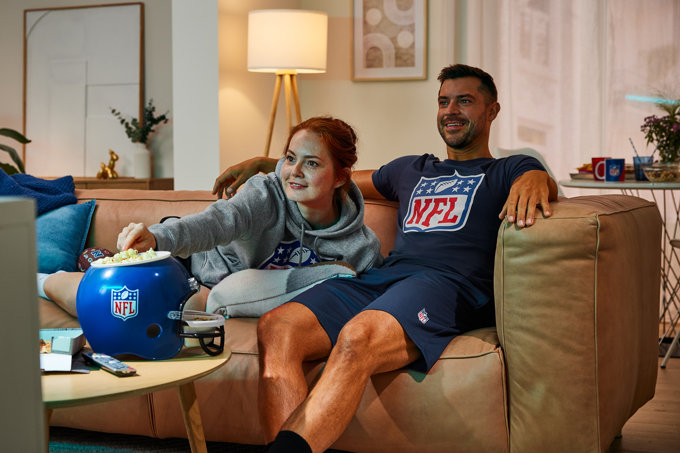 Man en vrouw kijken sport op tv, eten popcorn uit een helm en drinken uit mokken.