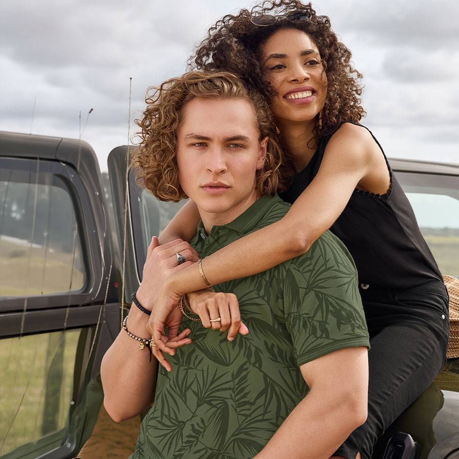 Lachend stel, hij in groen poloshirt met bladeren, zij in zwarte top, bij een auto.