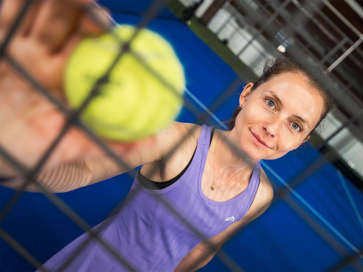Vrouw in sportkleding houdt padelbal door net, blauw veld op de achtergrond.