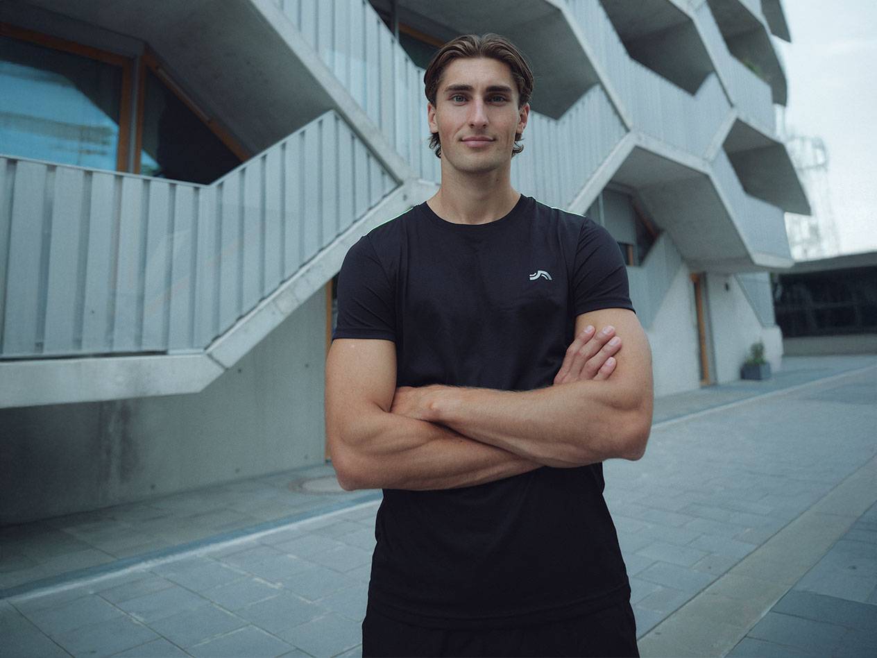 Man in zwart T-shirt met logo en zwarte korte broek, staat buiten met gekruiste armen.