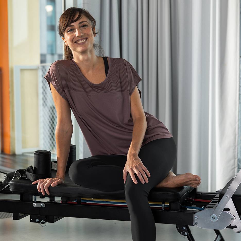 Vrouw in sportkleding lachend, zittend op een Pilates-apparaat.