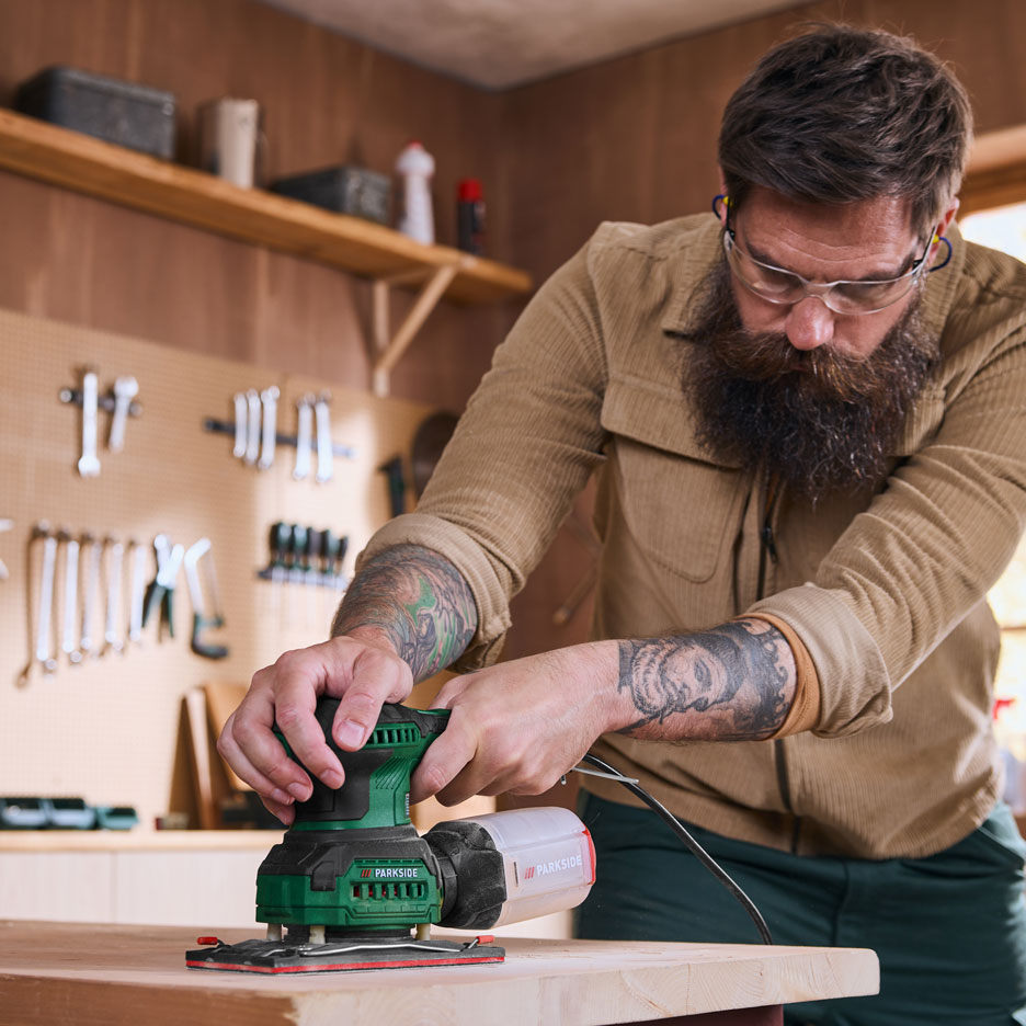 Man in werkplaats schuurt een houten plank met een elektrische schuurmachine.