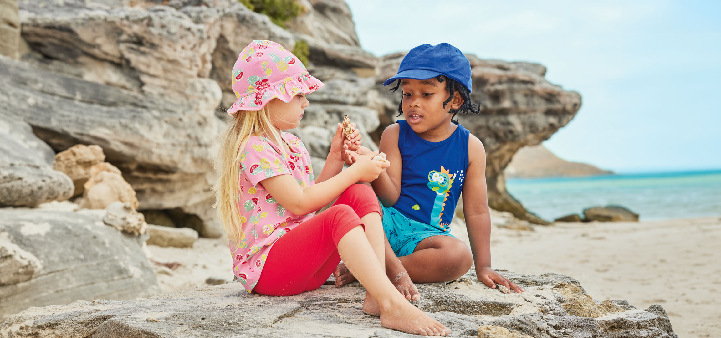Twee kinderen in zomerkleding zitten op een rots bij het strand, met een schelp.