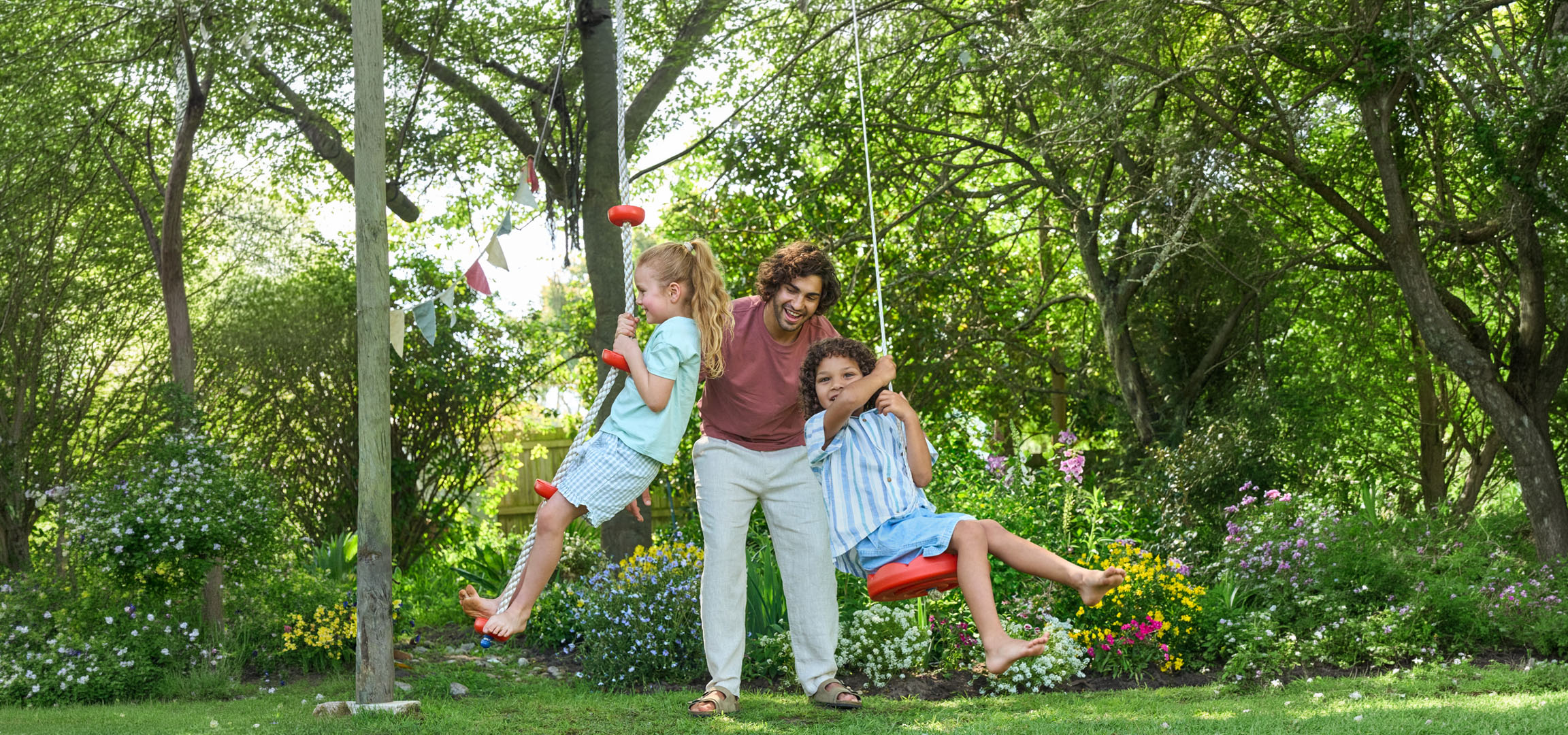 Vader en twee kinderen spelen op schommels in een weelderige groene tuin.