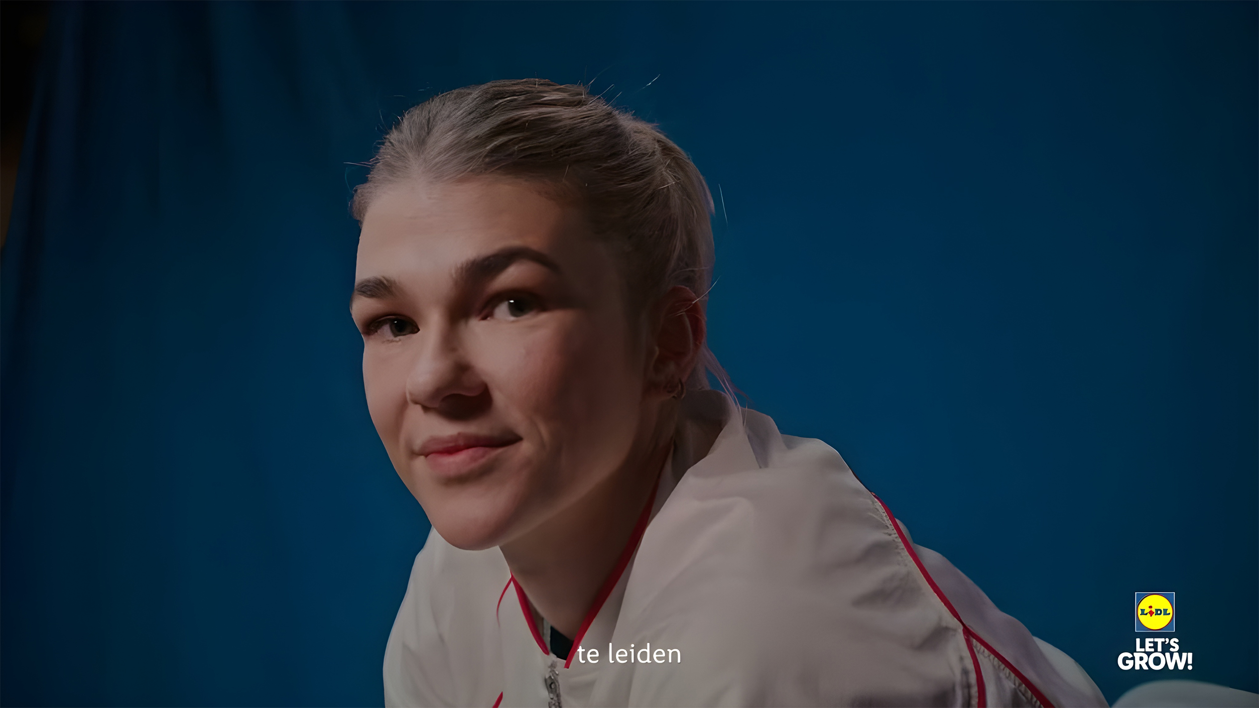 Een jonge vrouw in een wit sportjack met rode accenten, glimlachend, met het Lidl-logo en 'LET'S GROW!'.