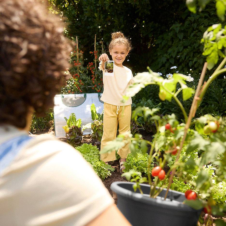 Kind in een tuin met een foto, met verse producten en een tomatenplant op de voorgrond.