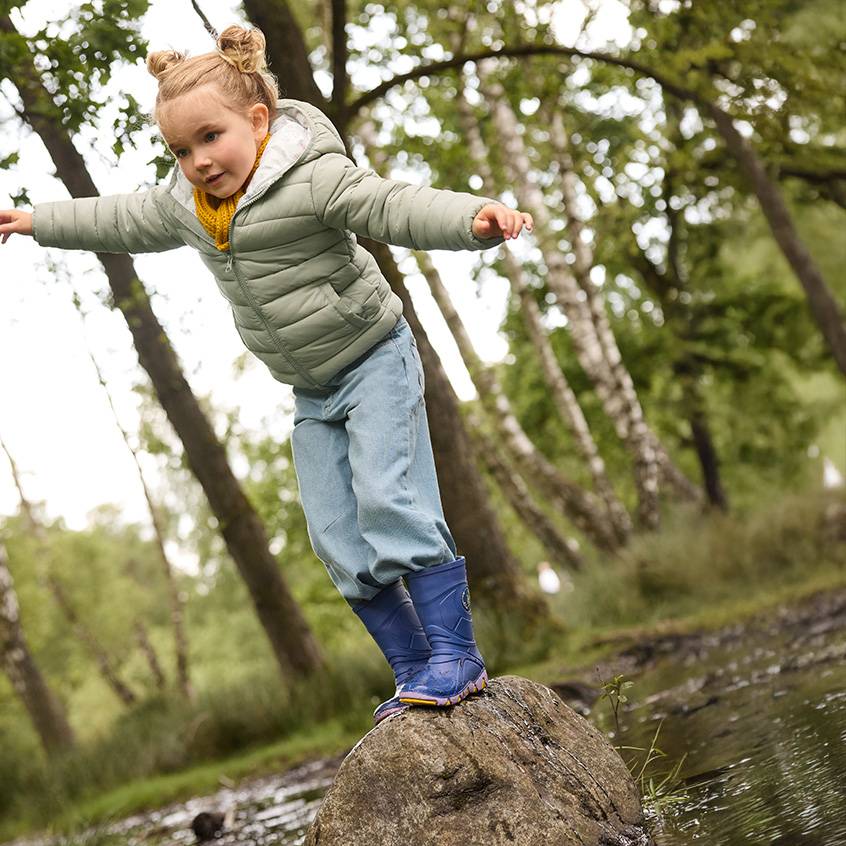 Klein meisje in een gewatteerde jas, jeans en regenlaarzen balanceert op een rots in het bos.