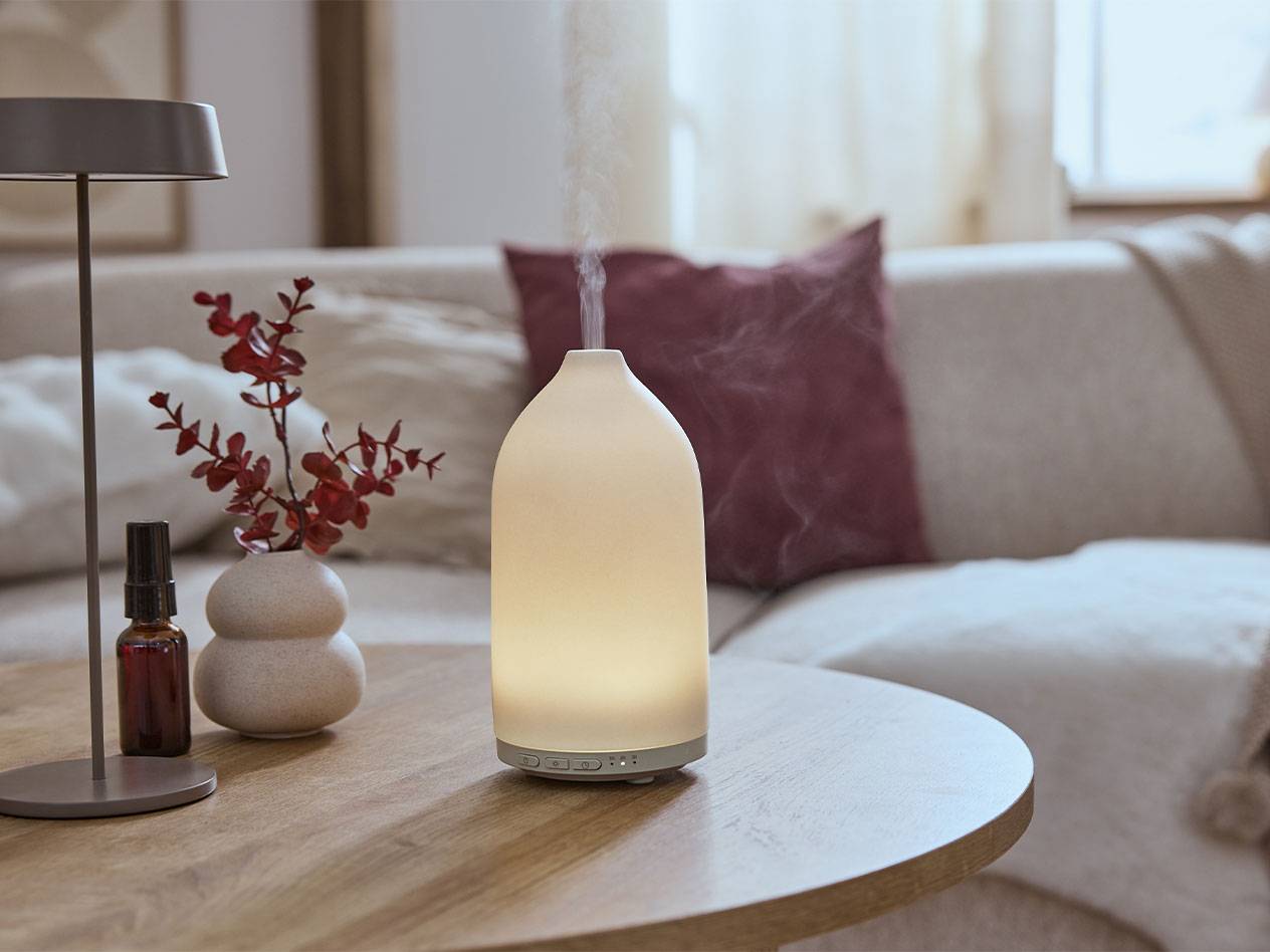 Verlichte aromadiffuser, oliefles en vaas met takken op een tafel.