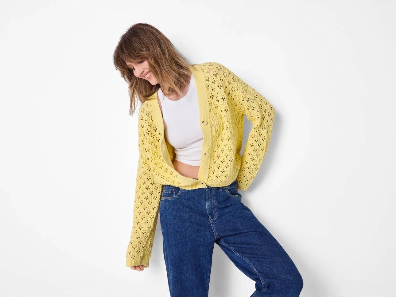 Vrolijke vrouw in gele gebreide cardigan, witte top en blauwe jeans.