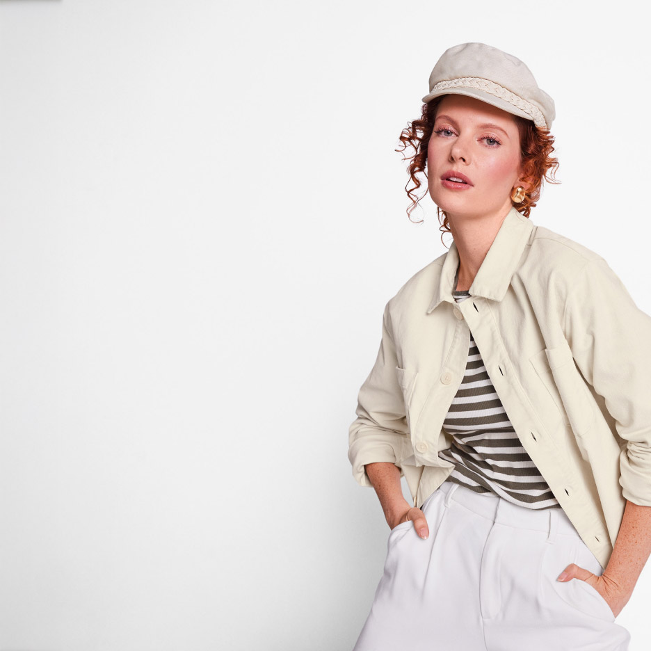 Vrouw met krullend haar draagt een beige pet, gestreepte top, beige jas en witte broek.