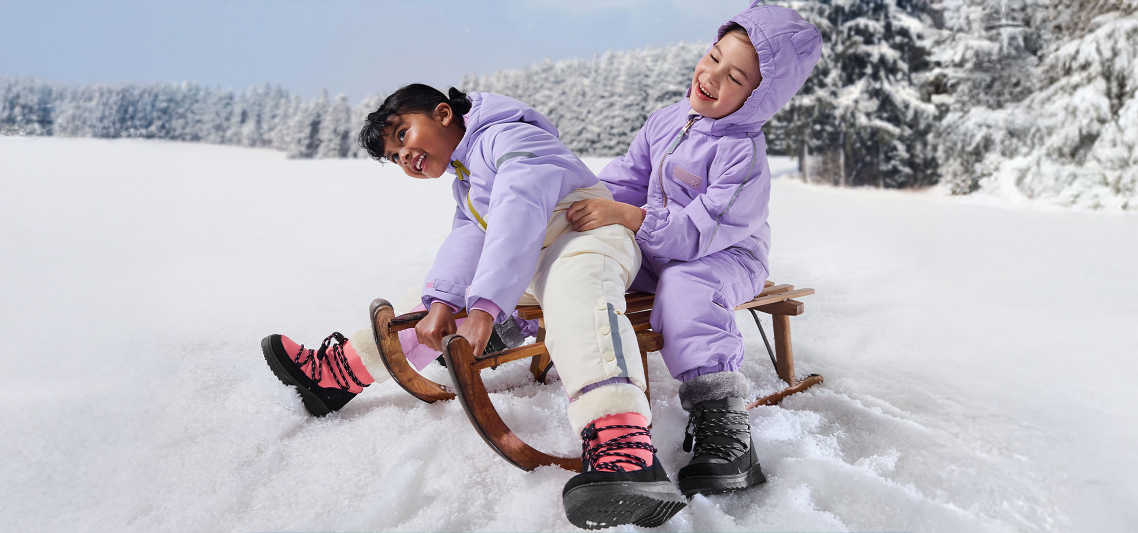 Twee kinderen in paarse skipakken en winterlaarzen sleeën in een besneeuwd landschap.