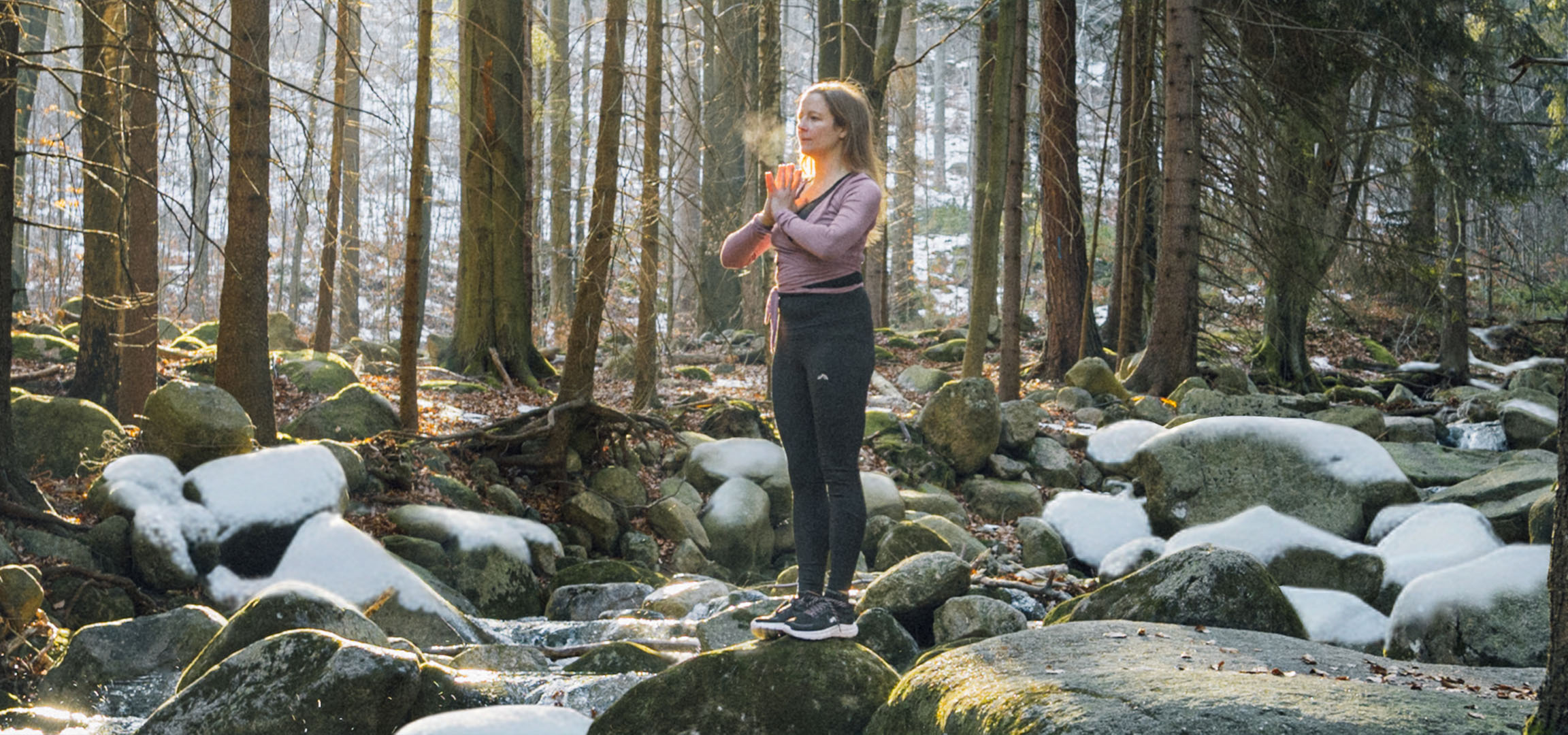 Vrouw in sportkleding die yoga doet in een besneeuwd bos met een beekje.