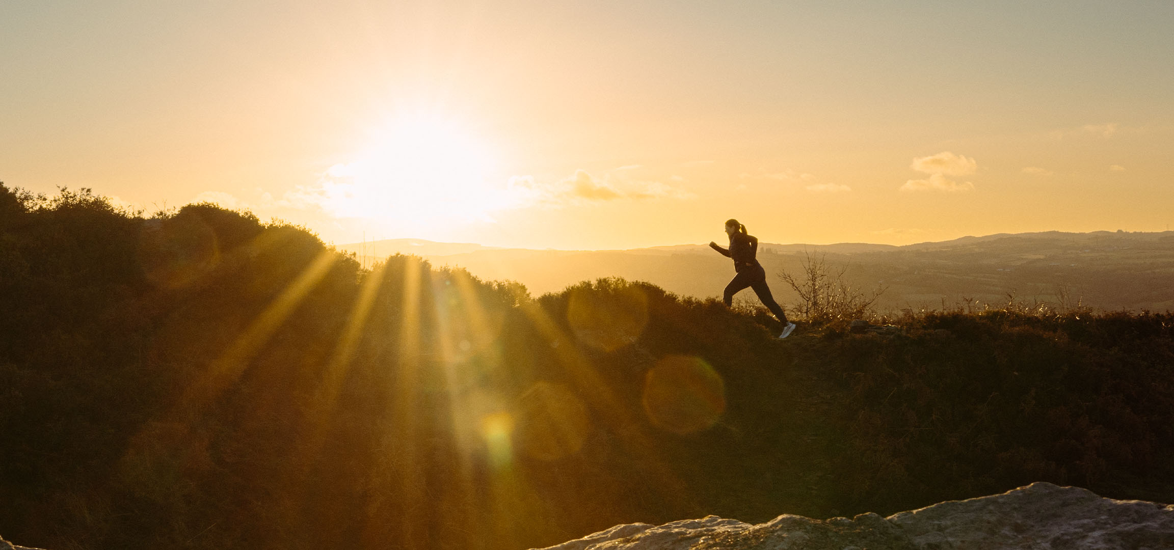 Vrouw rent op een heuvel bij zonsondergang, met zonnestralen en een schilderachtig landschap.