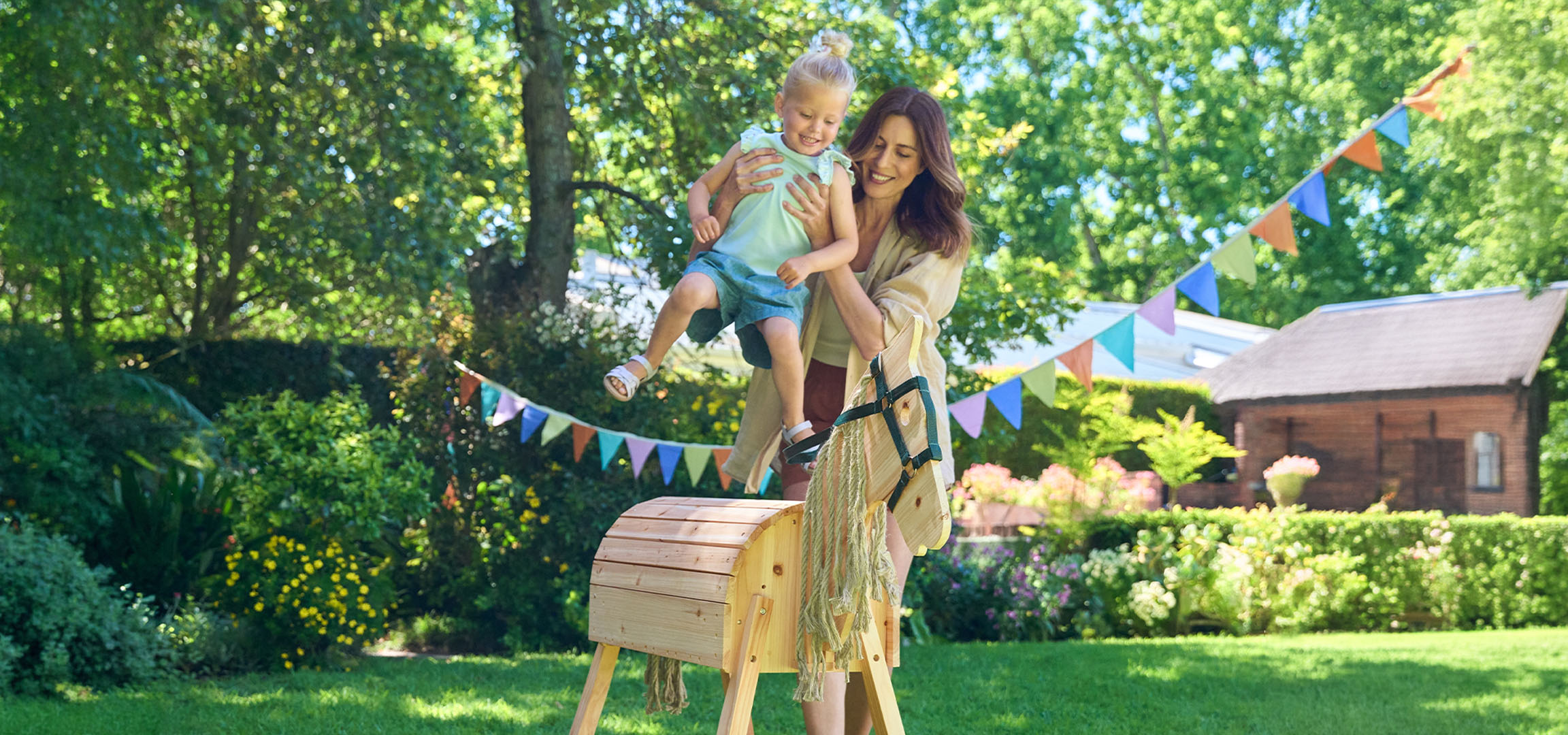 Moeder en kind spelen met een houten hobbelpaard in een zonnige tuin.