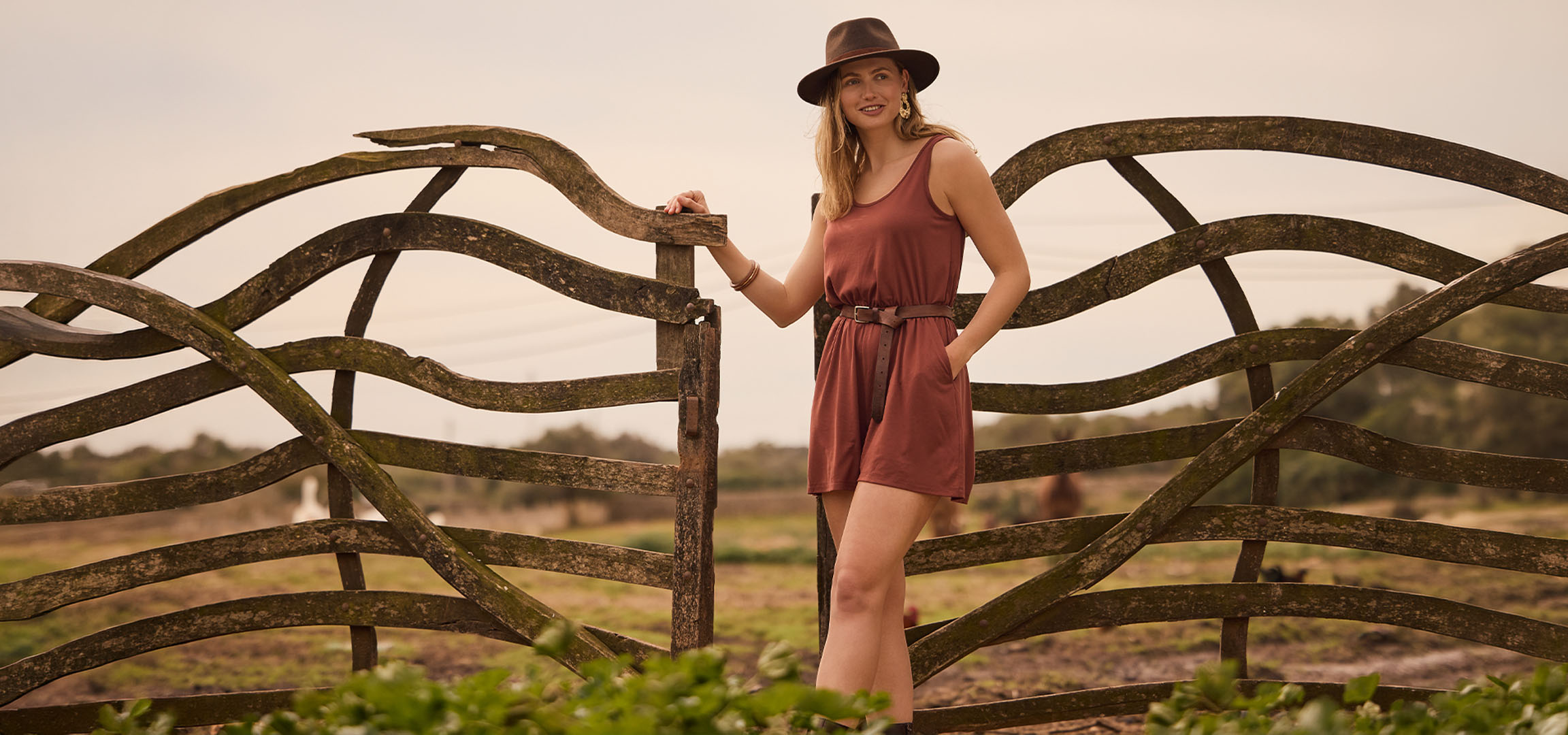 Vrouw in jurk en hoed, leunend tegen een houten hek op het platteland.
