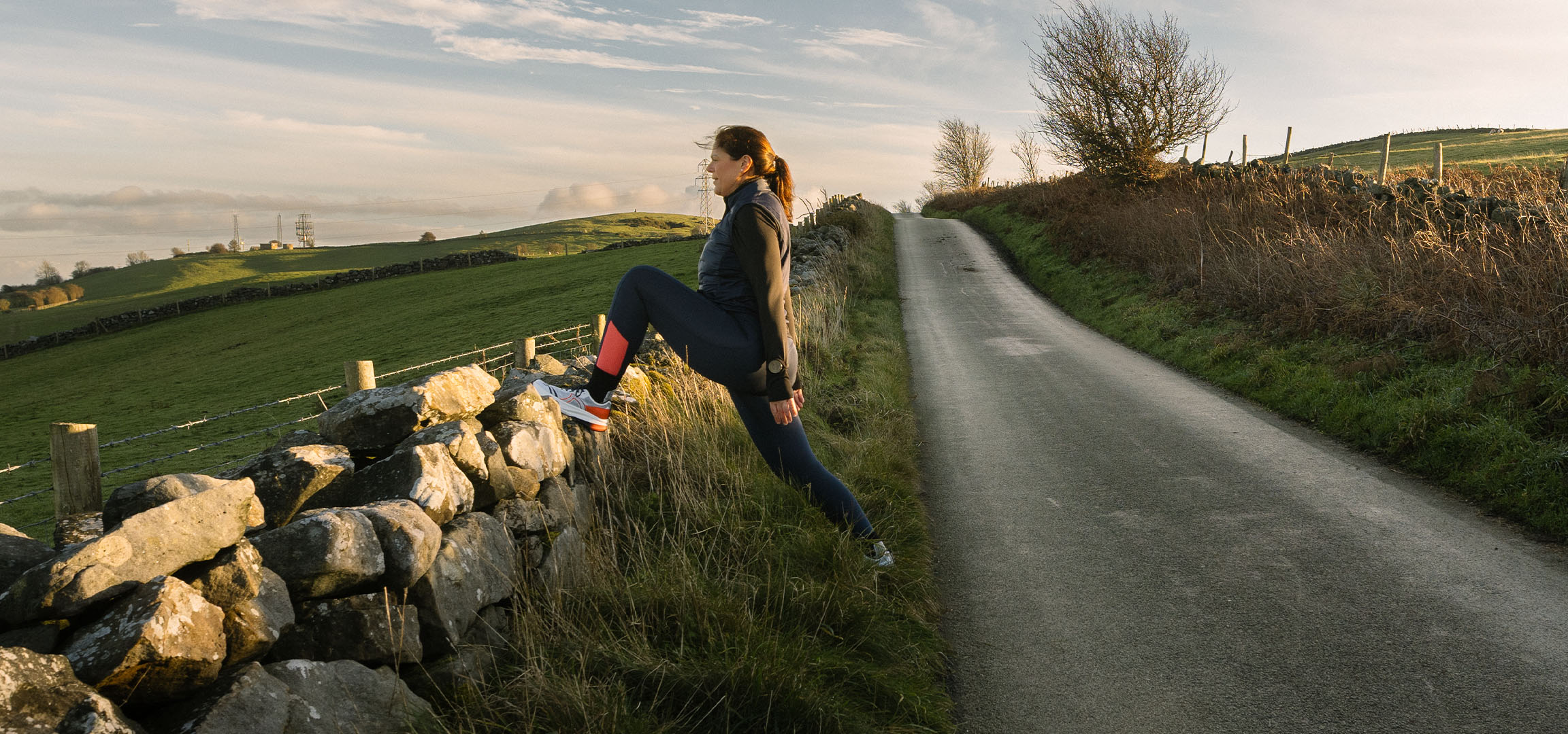 Vrouw rekt zich uit bij een stenen muur op een landweg bij zonsondergang.