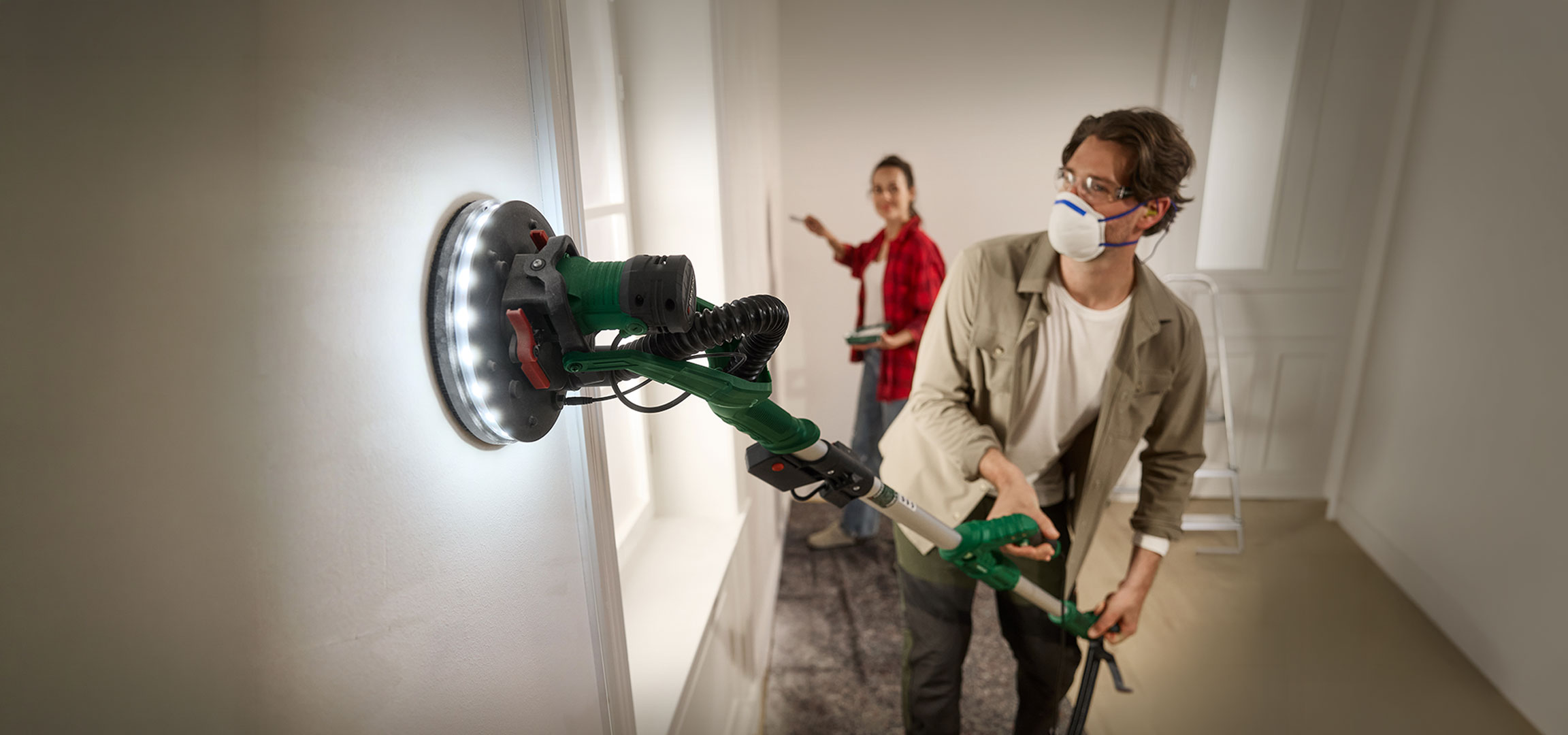 Gegenereerd door AI: Man schuurt muur met lange-hals schuurmachine, vrouw schildert op de achtergrond.