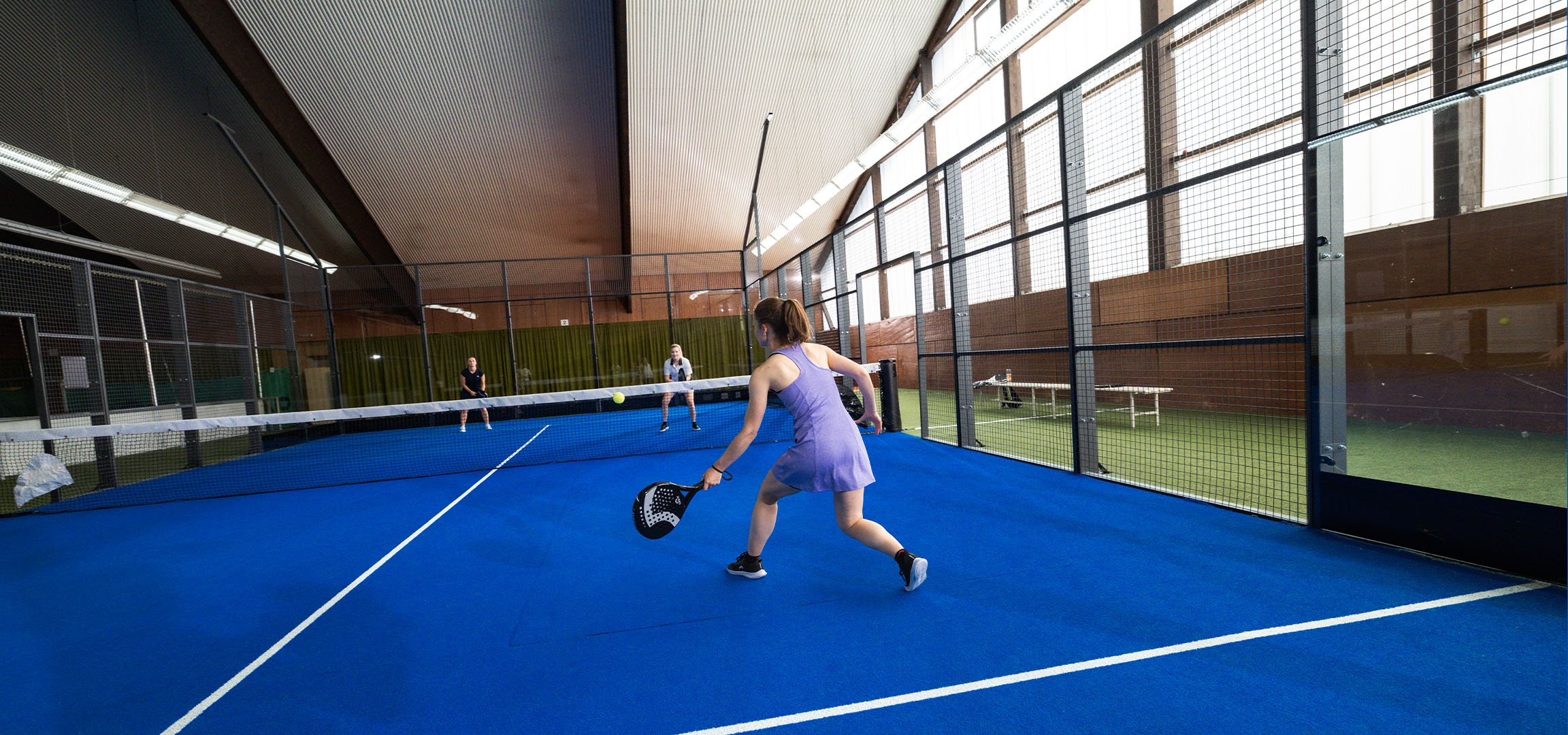 Vrouw speelt padel op een blauwe baan, met andere spelers op de achtergrond.